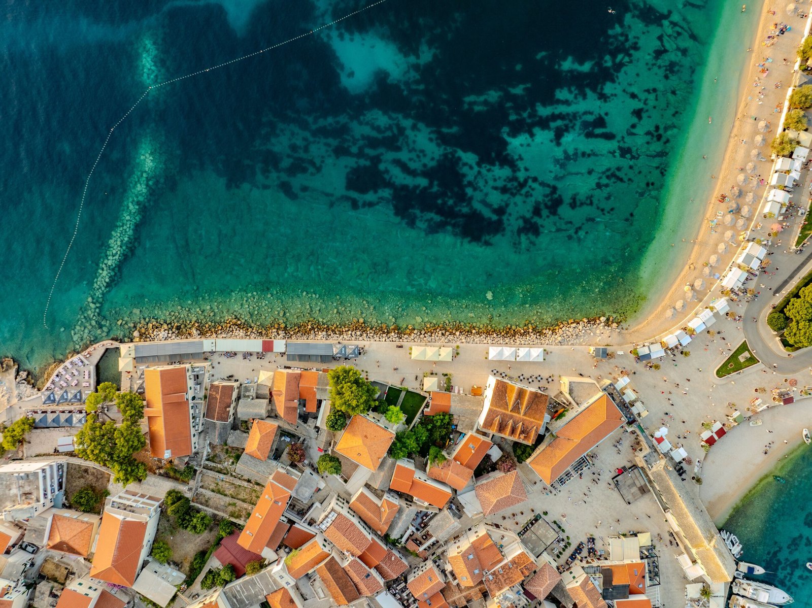 Aerial view of a coastal town with orange tile roofs, a rocky shoreline, and a crowded beach along turquoise water