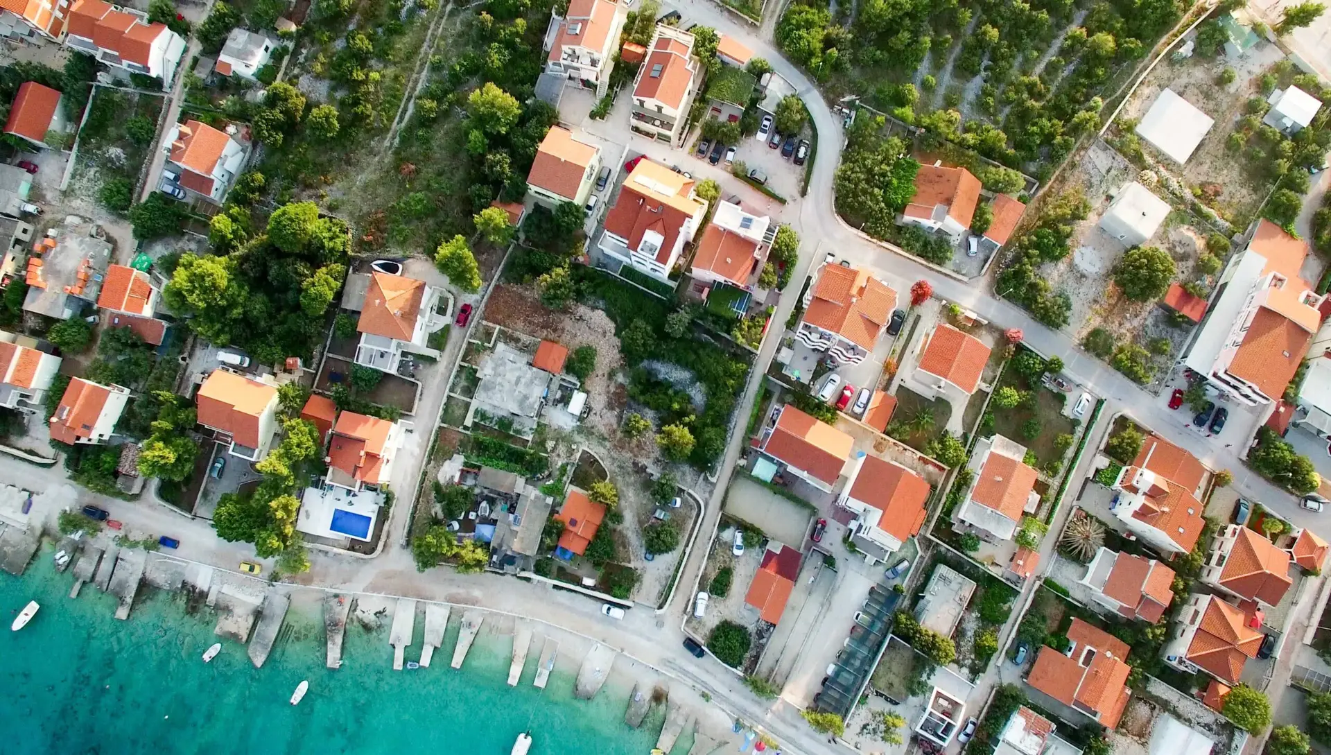 Aerial view of a coastal town with red-roofed houses, gardens, and small boats docked along turquoise water