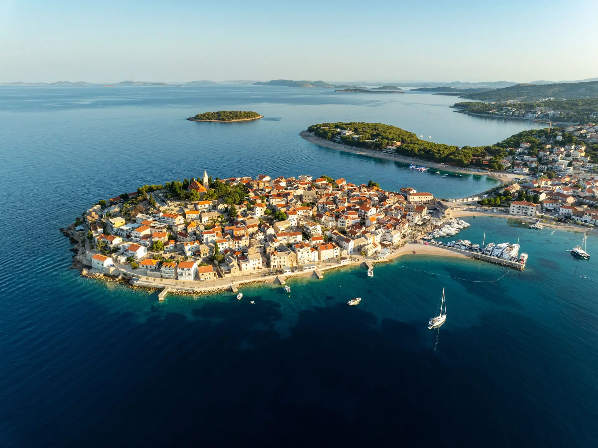 Aerial view of a coastal Croatian town on a peninsula with red-roofed buildings, surrounded by turquoise sea, boats, and nearby islands