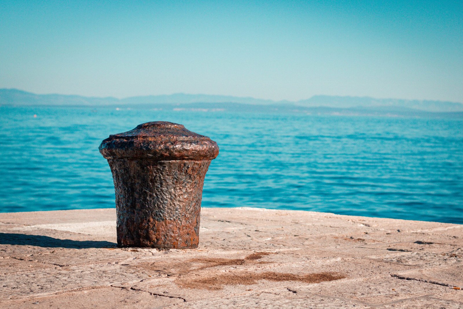 Rusty metal mooring bollard on a stone pier overlooking calm blue water and distant hills