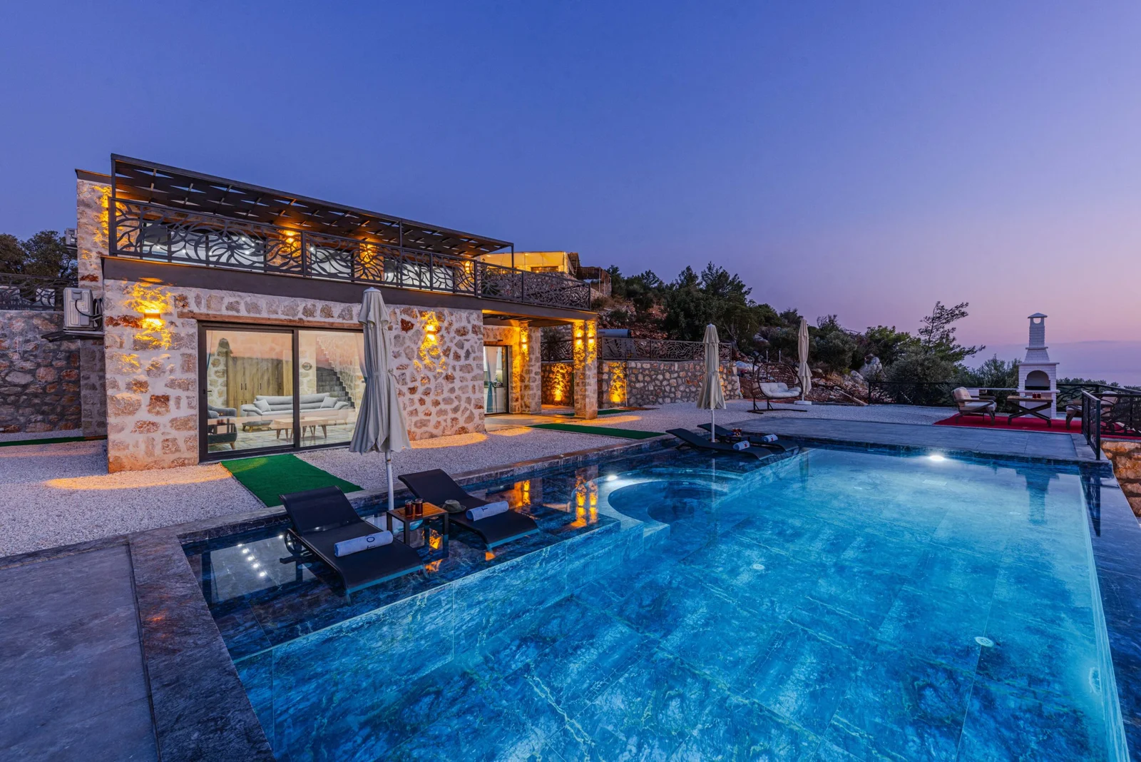 Stone villa with illuminated terrace and lit infinity pool with loungers at dusk, hills in background