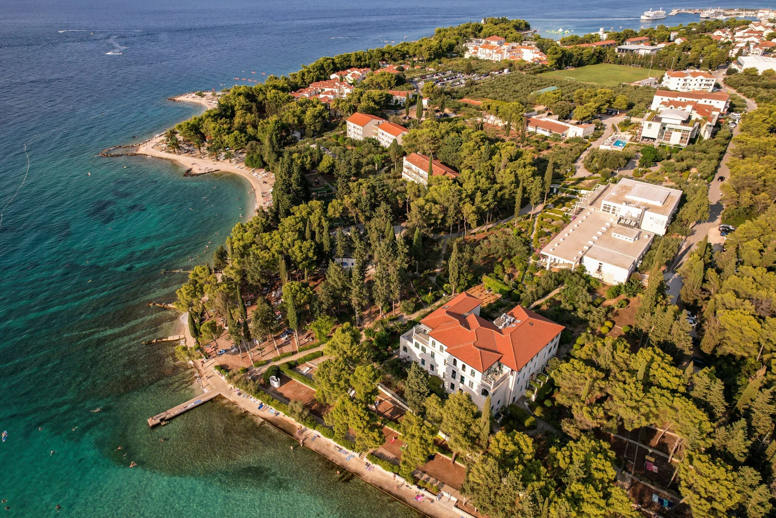 Aerial view of a wooded coastal resort peninsula with red-roofed villas, hotels, and beaches along turquoise waters